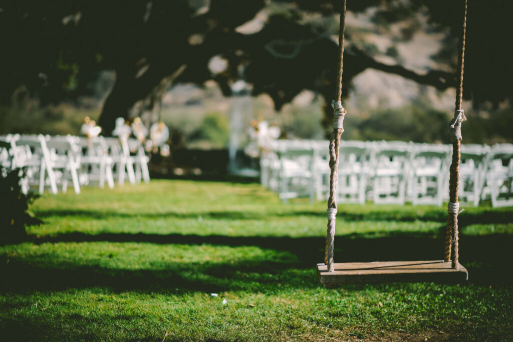 Sunlit grassy field with rustic wooden swing and white chairs for outdoor celebration under tree Wallpaper