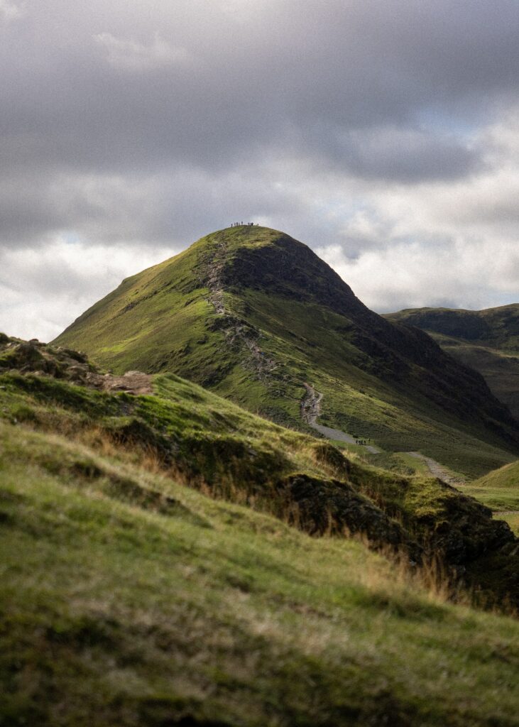 Lush green hill with winding path under dramatic clouds and sunlight highlighting summit silhouettes Wallpaper