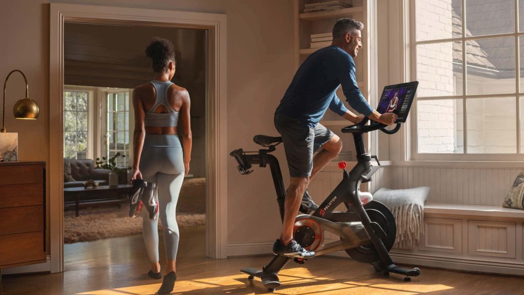 Man riding Peloton bike in sunlit modern home gym with woman holding cycling shoes nearby Wallpaper