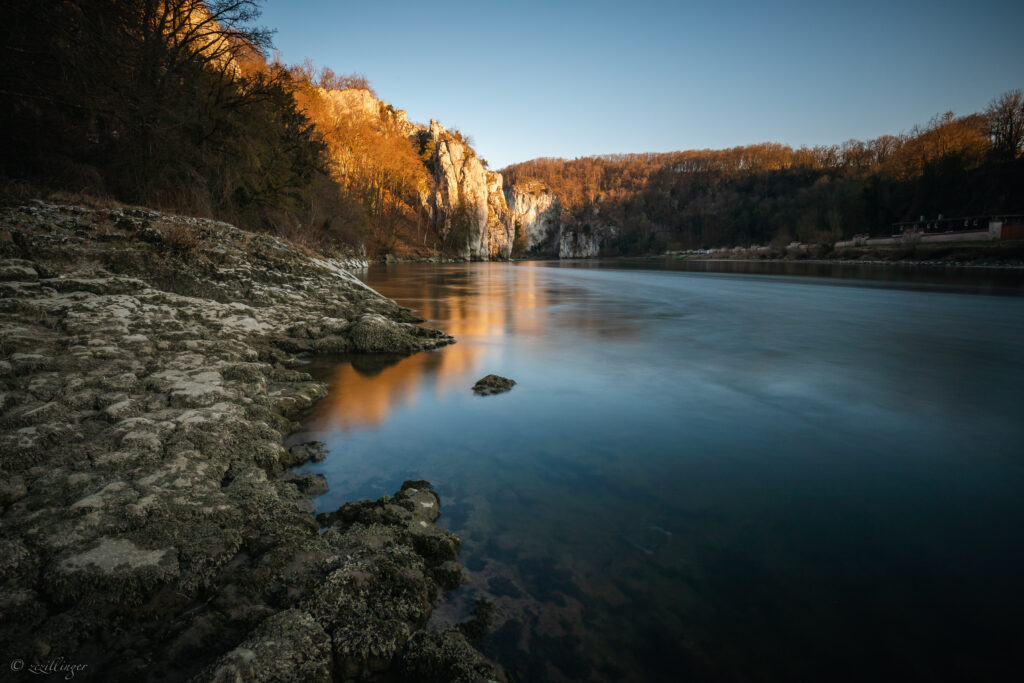 Serene lakeshore sunset with rugged rocks calm water sunlit cliffs and dense forest wallpaper background