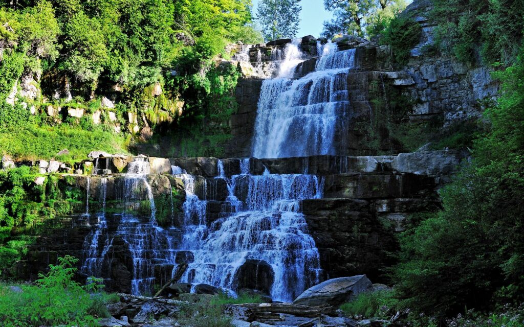 Multi-tiered waterfall over rugged rocks with lush green grass and dense summer foliage Wallpaper