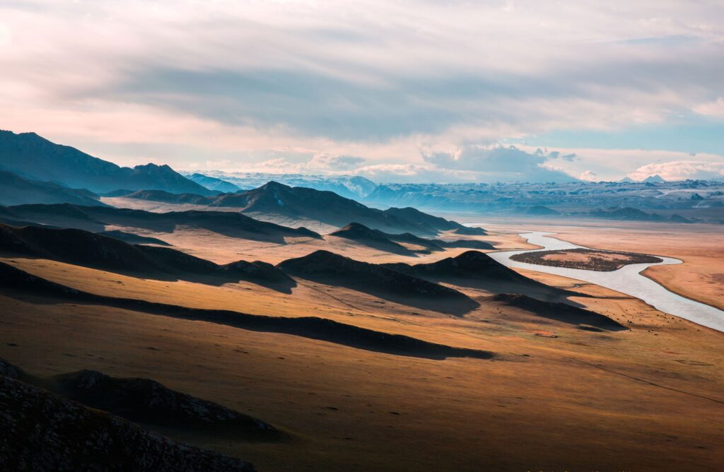 Prairie landscape with winding highway and river toward mountains under soft clouds sunlight Wallpaper