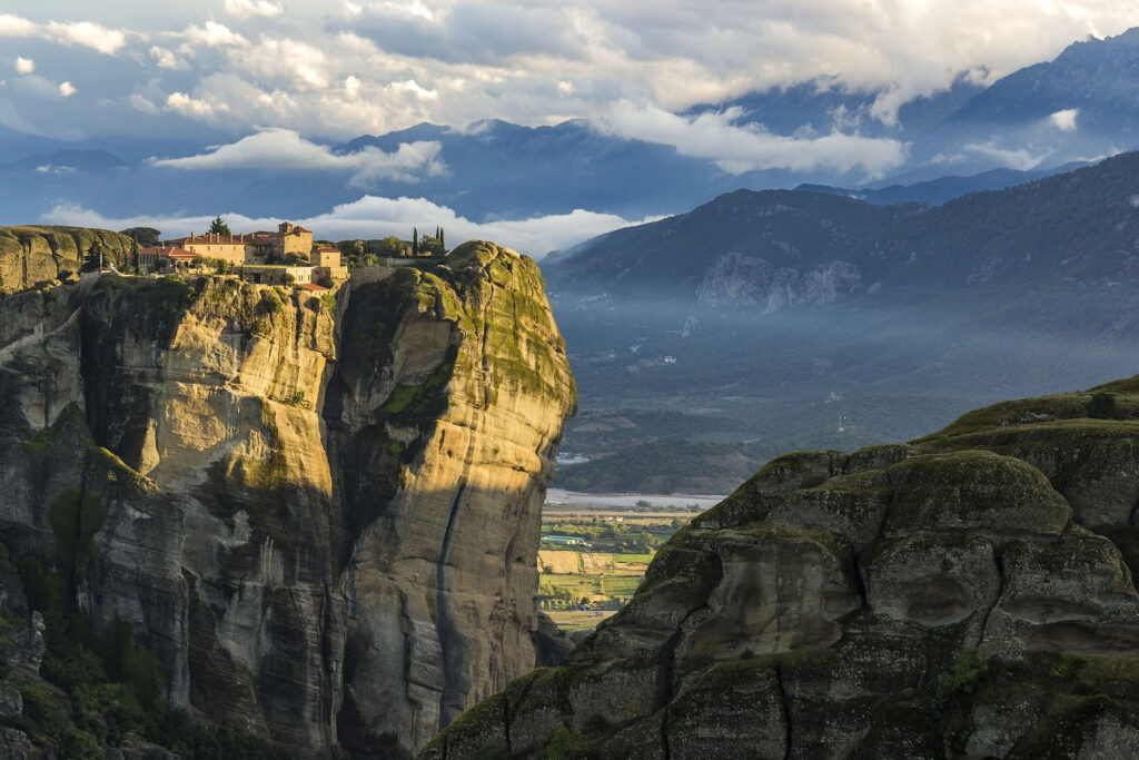 Sunlit monastery atop rugged cliffs with lush valley and rolling mountains under cloudy sky wallpaper