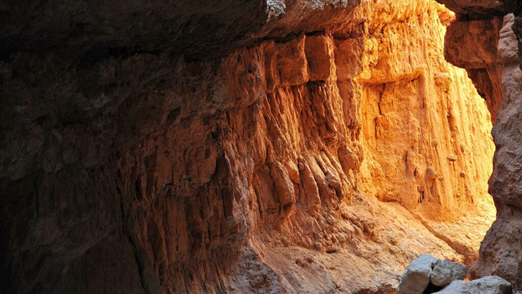 Sunlit layered rock cave walls with rich earthy tones and dramatic light and shadow textures Wallpaper