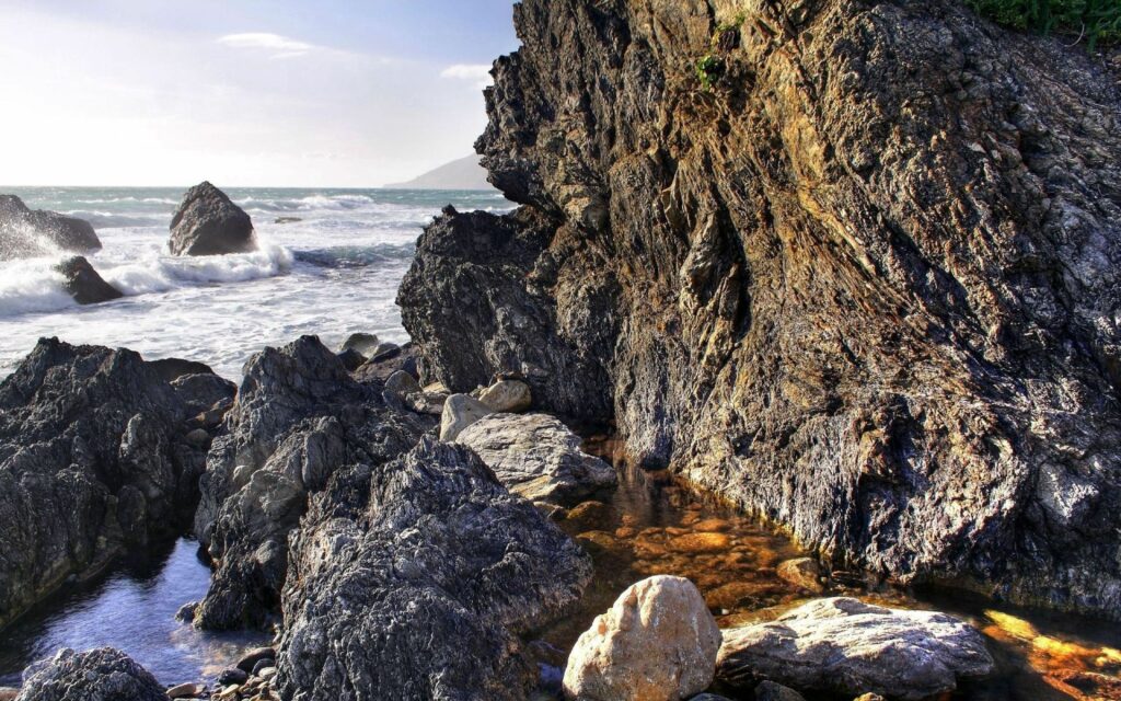 Sunlit rugged coastal rocks with waves crashing vibrant blue sea dramatic seascape wallpaper
