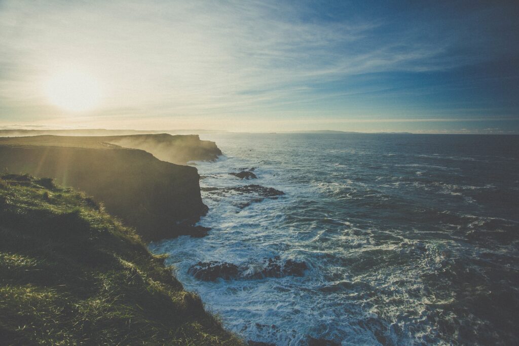 Sunlit rugged cliffs rise above misty waves and vibrant green coastline under a clear blue sky Wallpaper
