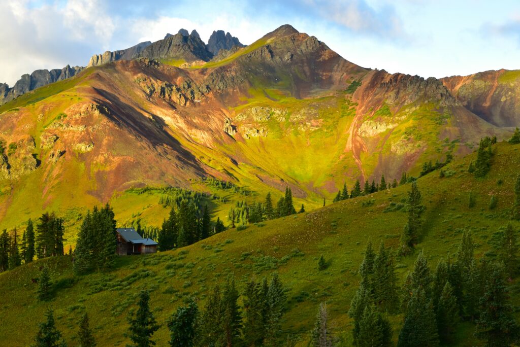Sunlit rugged mountains with green forested hills and a solitary building in nature wallpaper