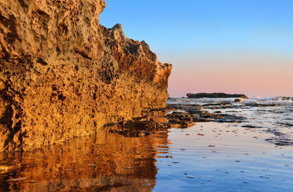 Rugged sunlit rock formation by calm sea with textured reflections under soft blue peach sky wallpaper