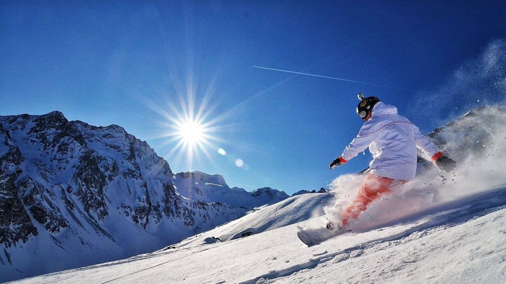 Snowboarder in white jacket and orange pants carving sunlit snowy mountain slope winter wallpaper
