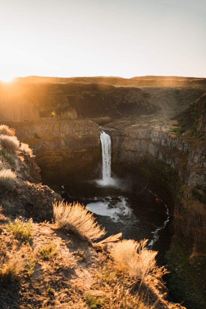 Waterfall cascading over rugged rocks and cliff under warm sunlight against clear blue sky wallpaper