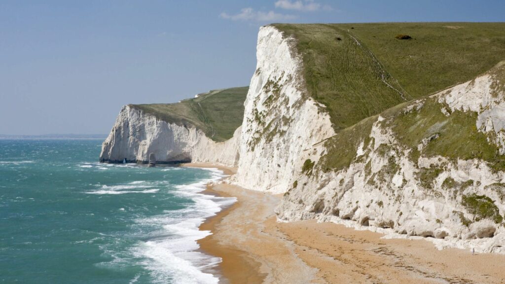 Dramatic white cliffs with rolling green hills above turquoise ocean waves and golden sandy beach Wallpaper