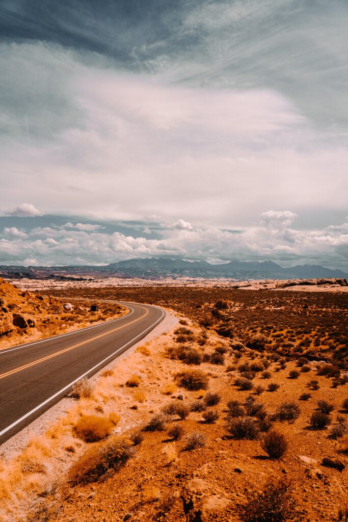 Winding road through sunlit prairie with dry bushes toward mountains under dramatic clouds Wallpaper