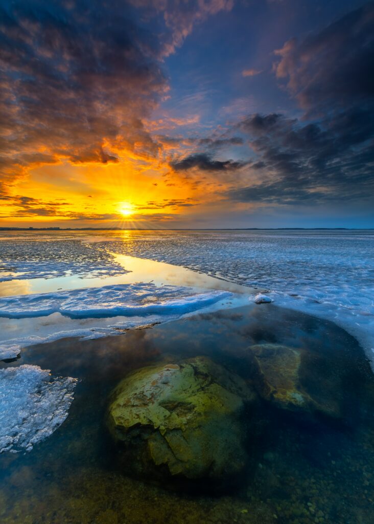 Sunrise over frozen horizon with orange blue sky reflecting on icy water and large stones Wallpaper