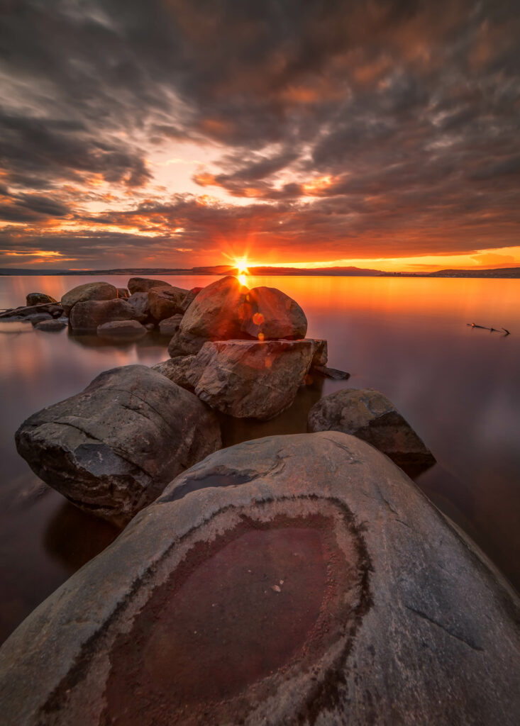 Sunrise over smooth lake waters with rugged stones and dramatic clouds in a golden sky Wallpaper