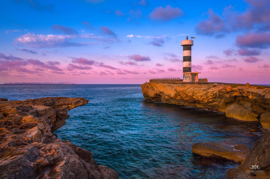 Striped lighthouse on rugged rocks by calm bay in Spain at sunrise serene coastal wallpaper background