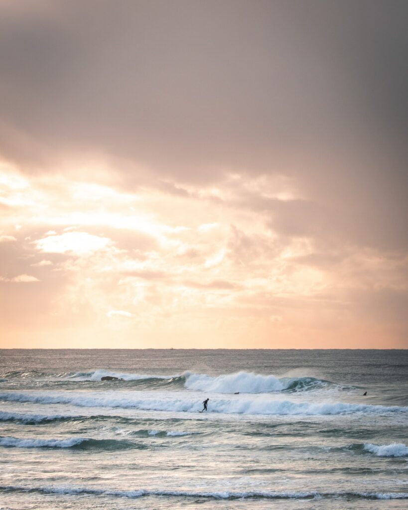 Surfer riding ocean waves at sunrise with pastel sky and golden light reflecting on water wallpaer Wallpaper