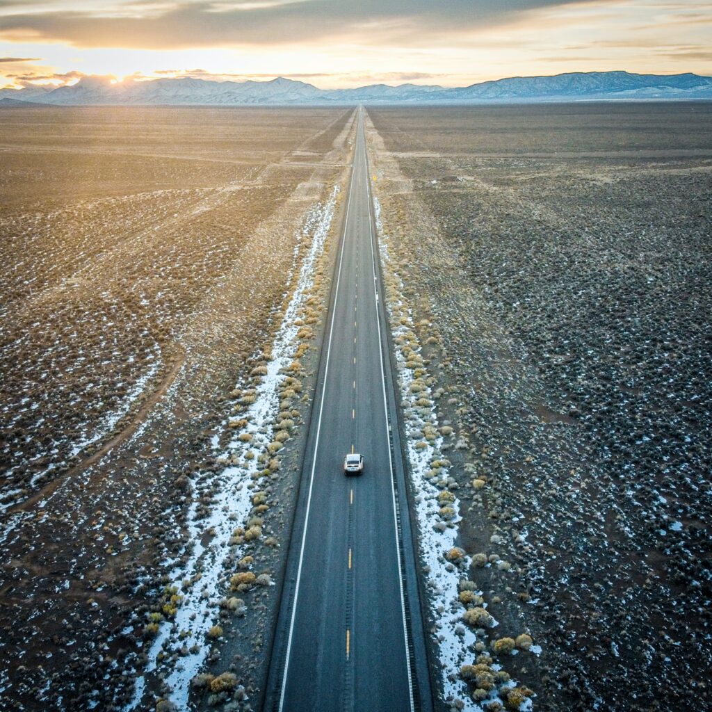 Aerial view of a lone car on straight road through rugged mountain valley at sunset with snow and desert Wallpaper