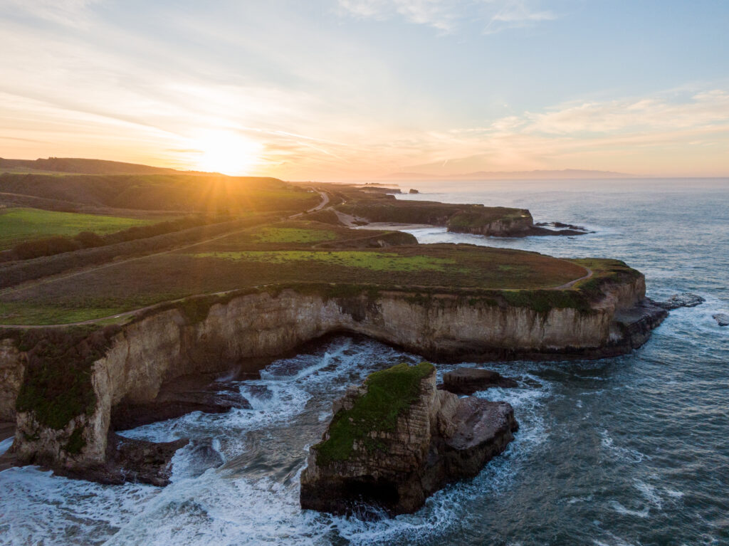 Aerial view of rocky coastline with cliffs lush greenery and colorful sunset over swirling sea Wallpaper