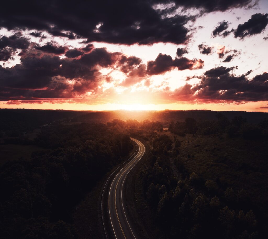 Aerial view of winding road through dense forest at sunset with orange and purple clouds sky Wallpaper