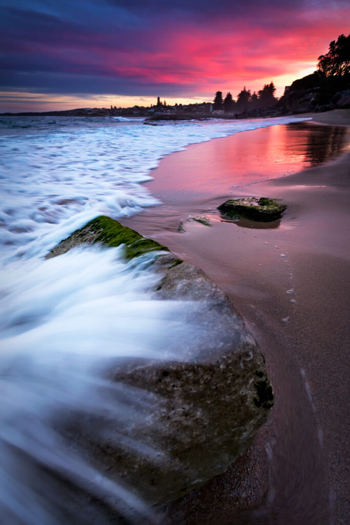 Sunset beach wallpaper with waves over rocks and sea foam on golden sand for relaxation