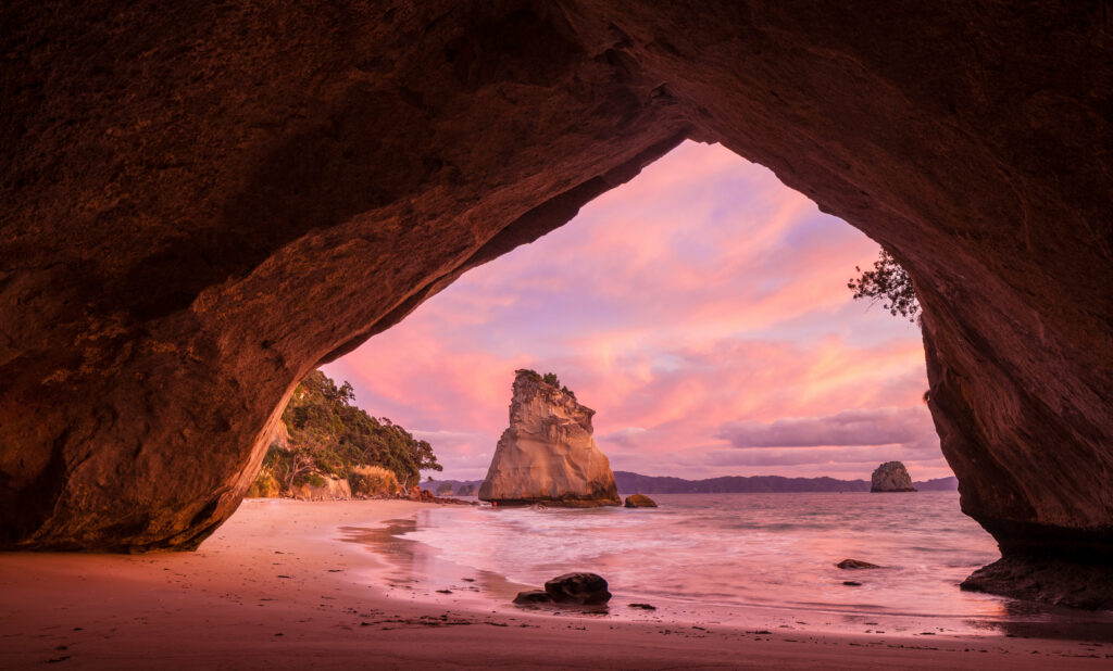 Beachscape wallpaper with natural rock cave framing sunset shoreline and sea stack over waves