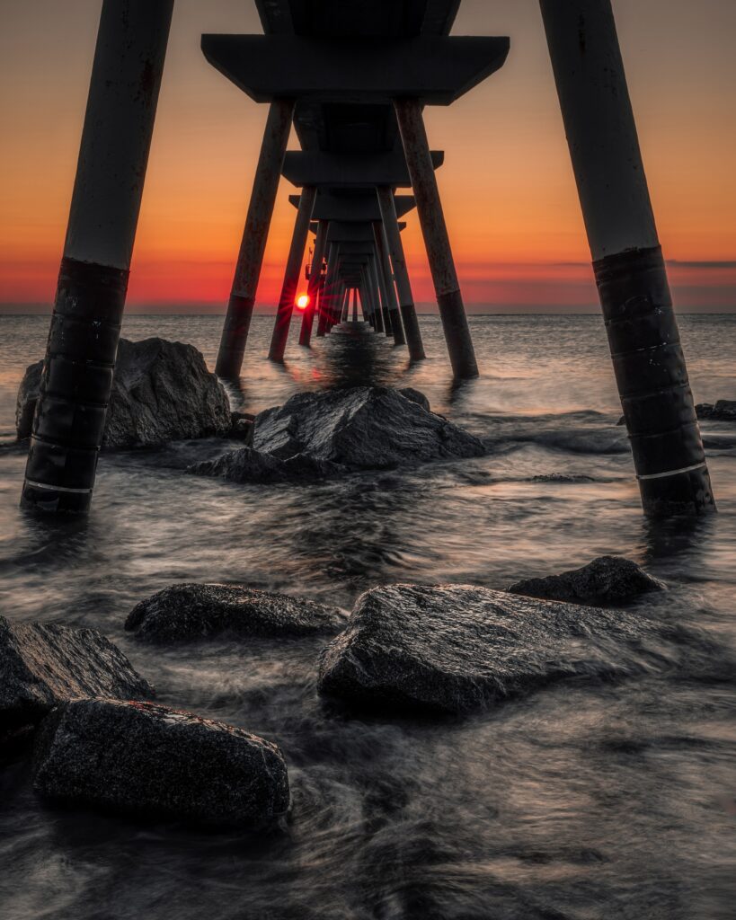 Sunset over seaside pier with orange sky reflecting on water and rugged stones foreground wallpaper