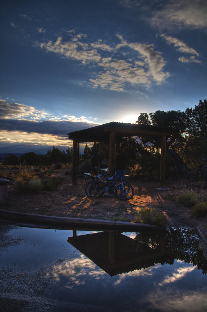 Two bicycles under rustic gazebo at Utah sunset with vibrant sky and reflective puddle background Wallpaper