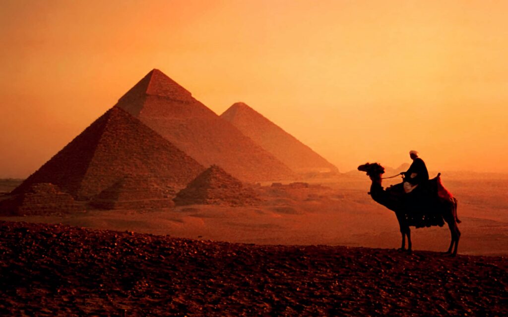 Person riding camel at sunset near pyramids in Egypt with warm orange tones and desert landscape Wallpaper