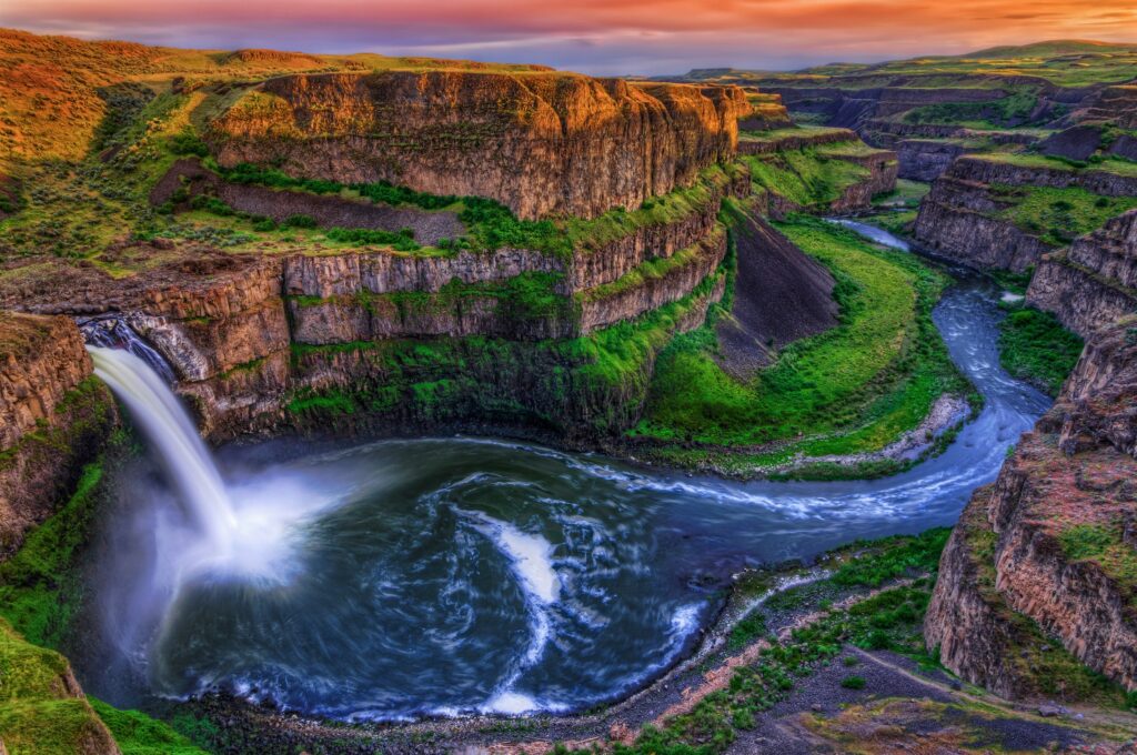 Sunset canyon waterfall plunges into swirling river surrounded by layered rocks and greenery Wallpaper