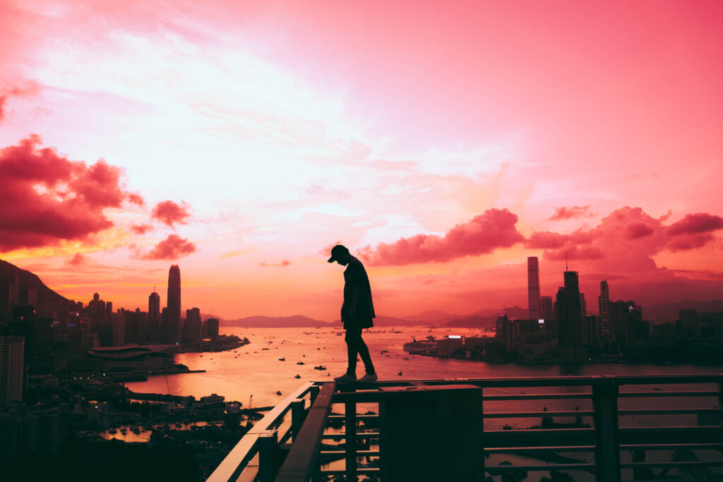 Man on rooftop at sunset overlooking city skyline with vibrant orange pink sky and calm water Wallpaper