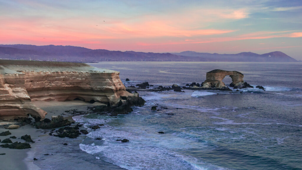 Coastal arch at sunset with rugged rocks gentle waves and distant mountains serene seaside view Wallpaper