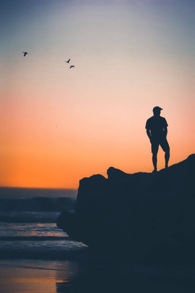 Silhouette of man in cap on rocky cliff at sunset ocean gradient sky peaceful wallpaper background