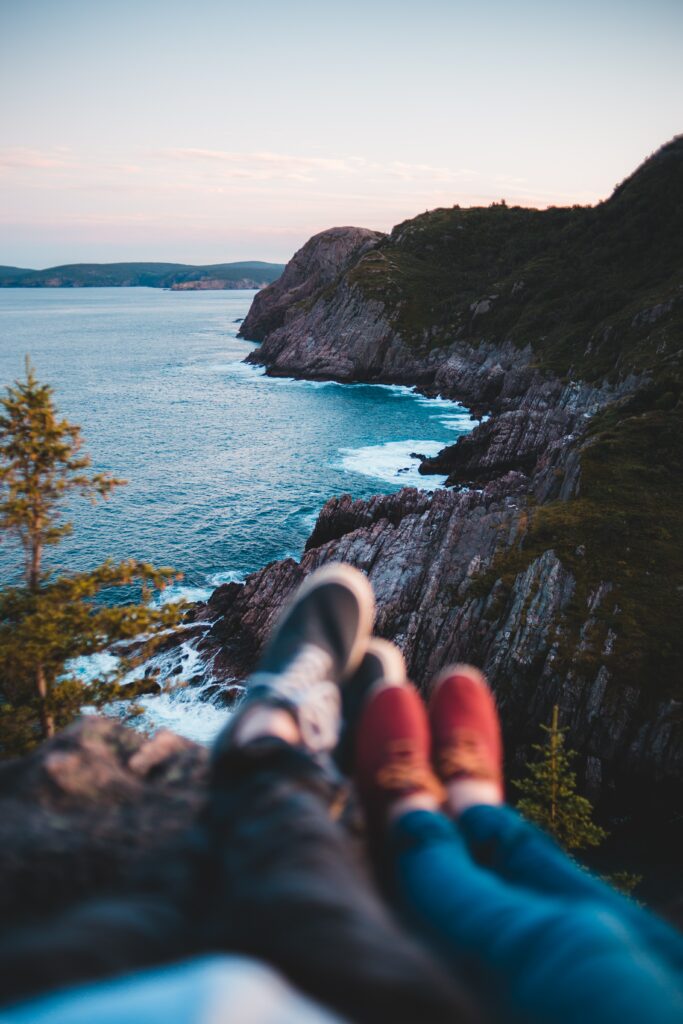 Two people sit on rocky cliff overlooking calm blue sea and lush shore trees at sunset wallpaper