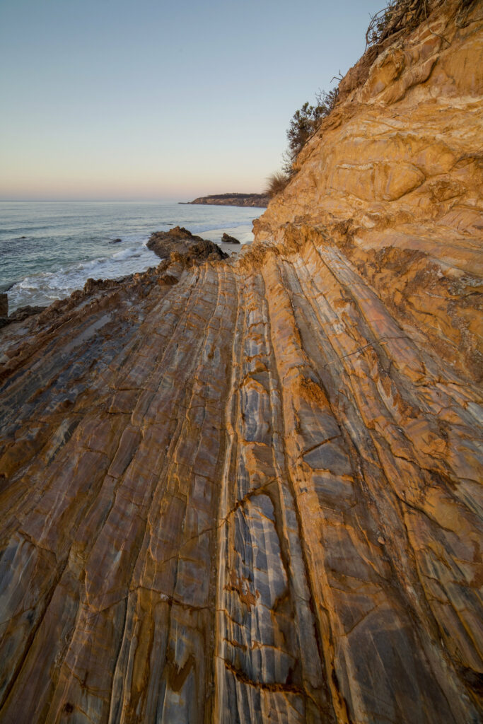 Sunset over rugged coastal cliff with layered rocks and calm sea under soft clear sky wallpaper
