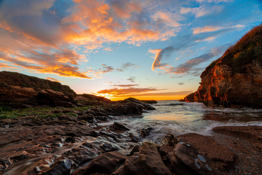 Sunset coastal landscape with rugged rocks gentle waves vibrant orange blue tones wallpaper