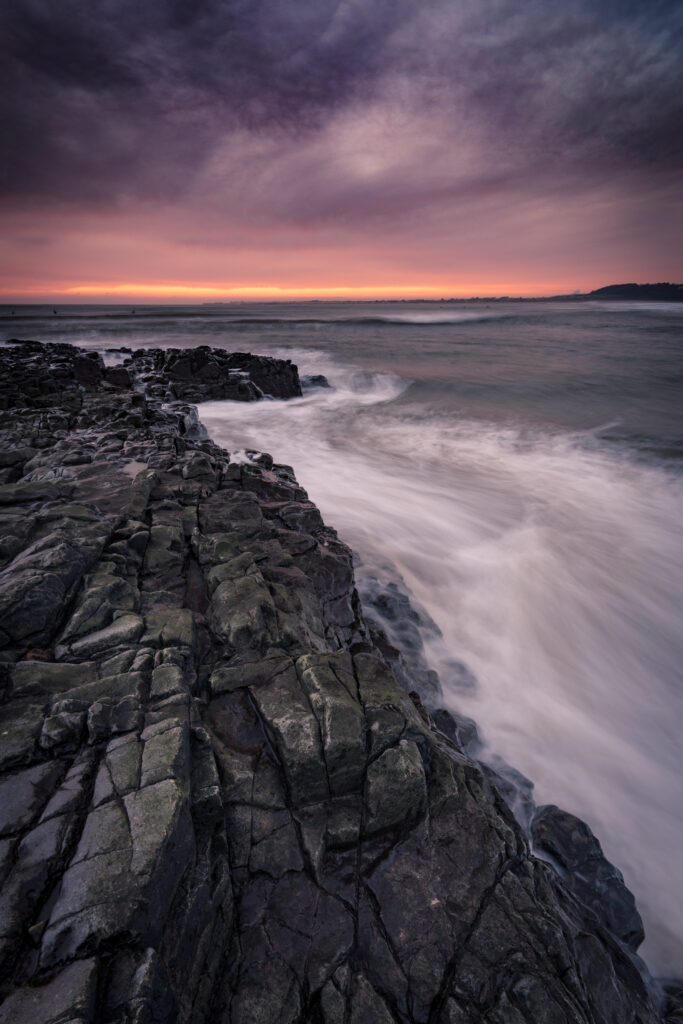 Dramatic coastal sunset with rugged rocks smooth stones crashing waves vibrant orange pink sky Wallpaper
