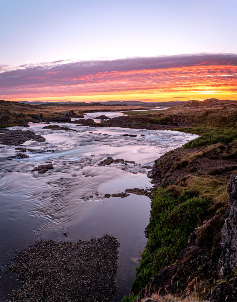 Coastal river at sunset with orange pink sky reflecting on calm water and lush green banks Wallpaper