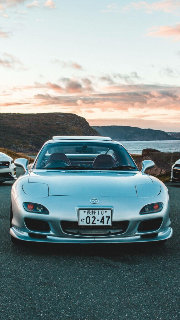 Silver Mazda RX-7 leads sleek sports cars parked on coastal road at sunset with cliffs ocean view Wallpaper