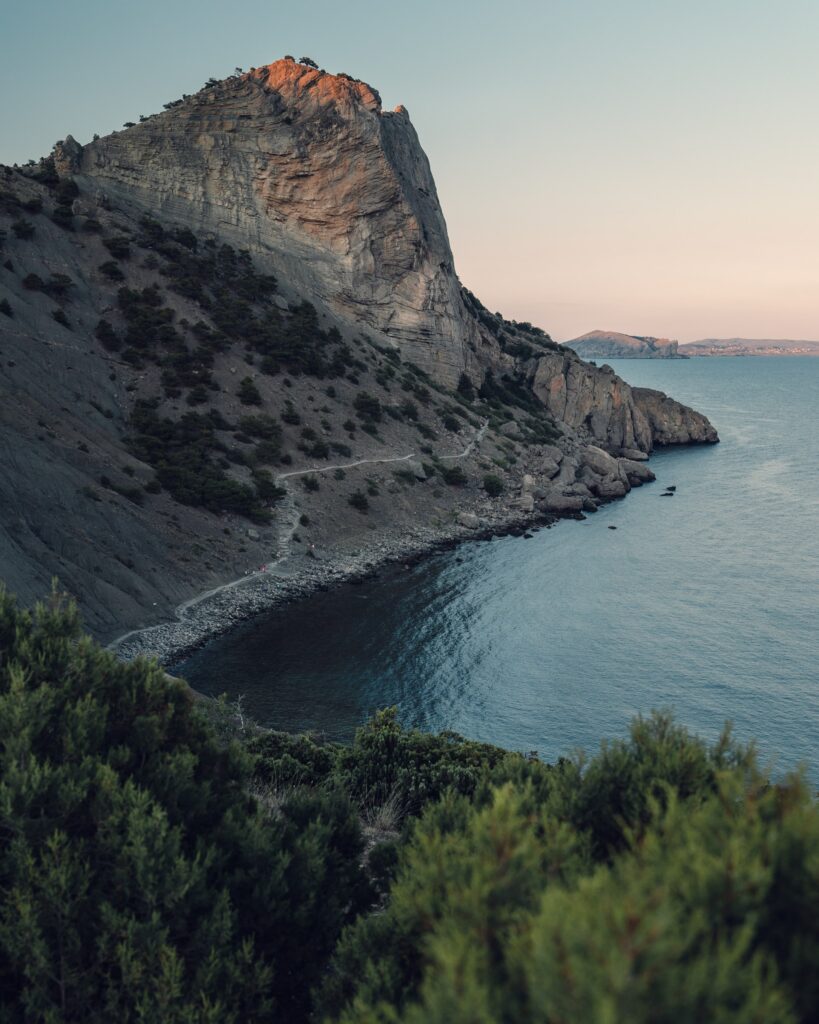 Dramatic coastal rock formation at sunset with rugged cliffs lush greenery and calm blue sea Wallpaper