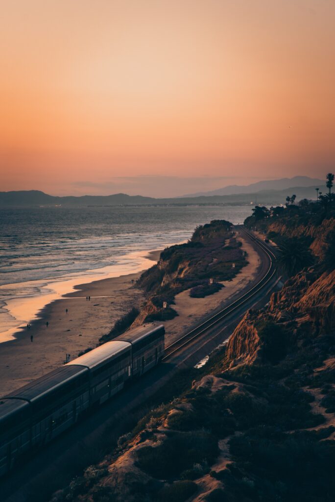 Sleek train travels coastal rails at sunset between sandy beach rugged cliffs and mountains Wallpaper