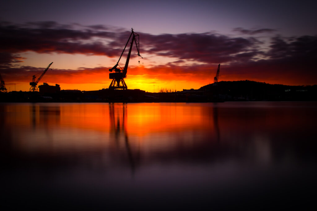 Sunset construction scene with towering cranes silhouetted against vibrant sky and water reflection Wallpaper