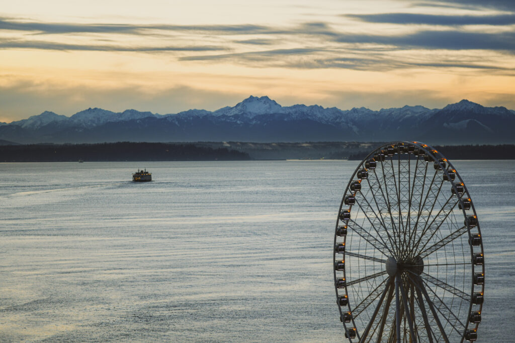 Colorful ferris wheel by sea at sunset with golden water reflections and distant mountains background Wallpaper