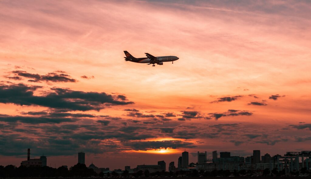 Commercial airplane flying at sunset over city skyline with orange pink sky and soft clouds Wallpaper
