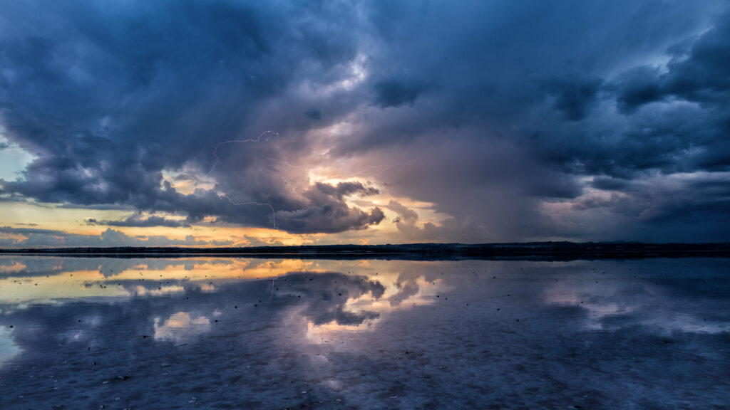 Dramatic sunset sky with towering clouds reflected on serene lake blending blues and golds Wallpaper