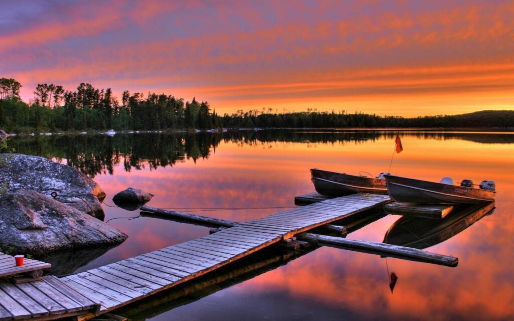 Two boats moored at rustic wooden pier on calm lake with vibrant sunset and tranquil trees Wallpaper