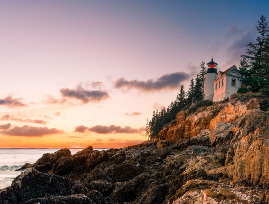 Lighthouse on rocky shore at sunset surrounded by pine trees and dramatic coastal clouds Wallpaper