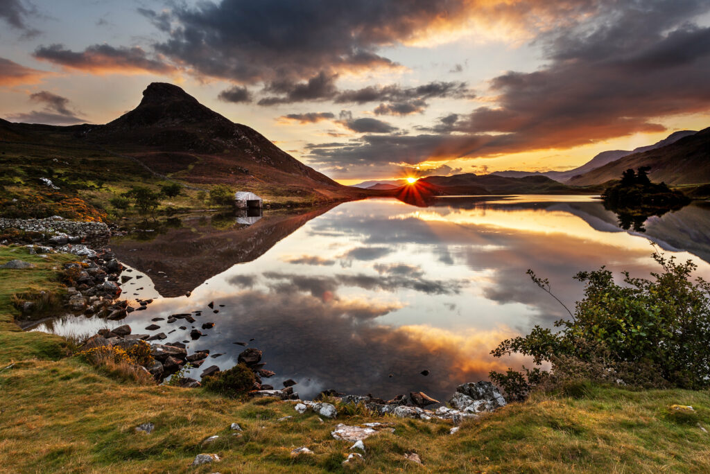 Sunset over rugged mountain and rocky shore reflected in calm lake with vibrant orange purple hues Wallpaper