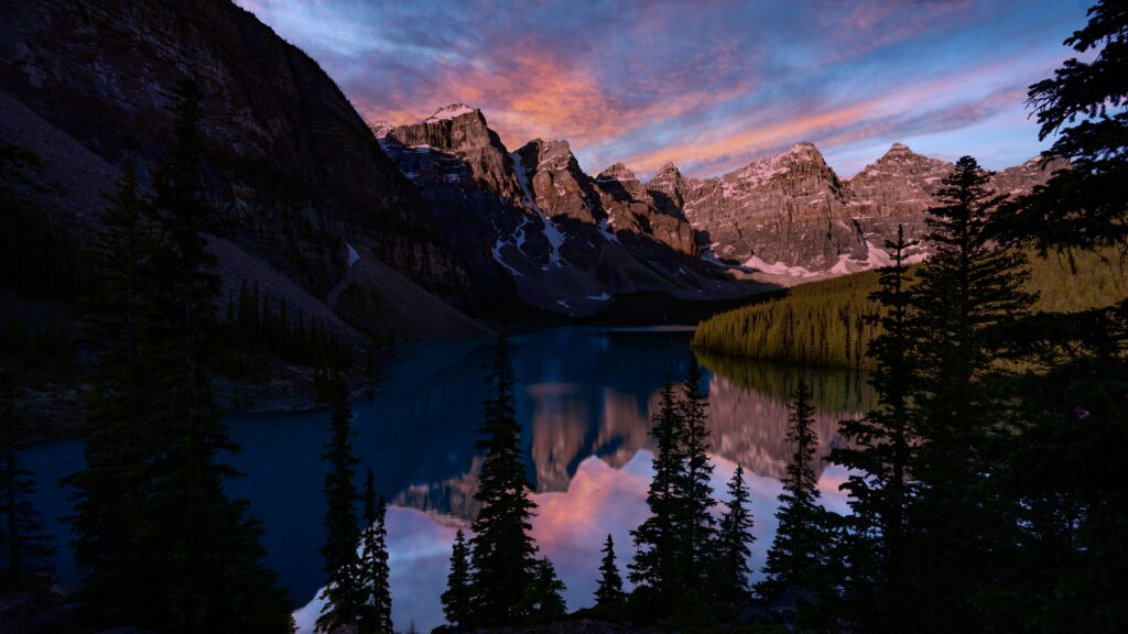 Sunset-lit mountains over a serene blue lake with evergreen trees in a tranquil landscape wallpaper