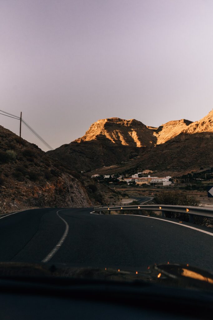 Winding asphalt road through rugged mountains at sunset with distant buildings in valley background Wallpaper