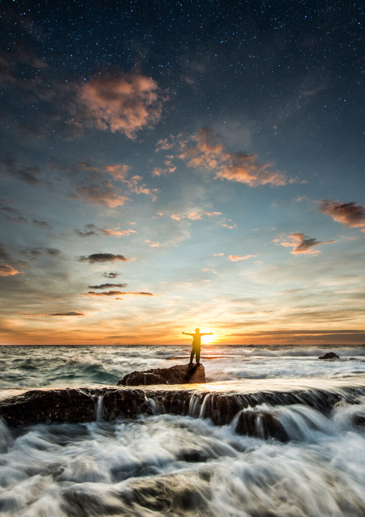 Silhouette of man on rocks by crashing ocean waves at sunset with golden light and starry sky Wallpaper
